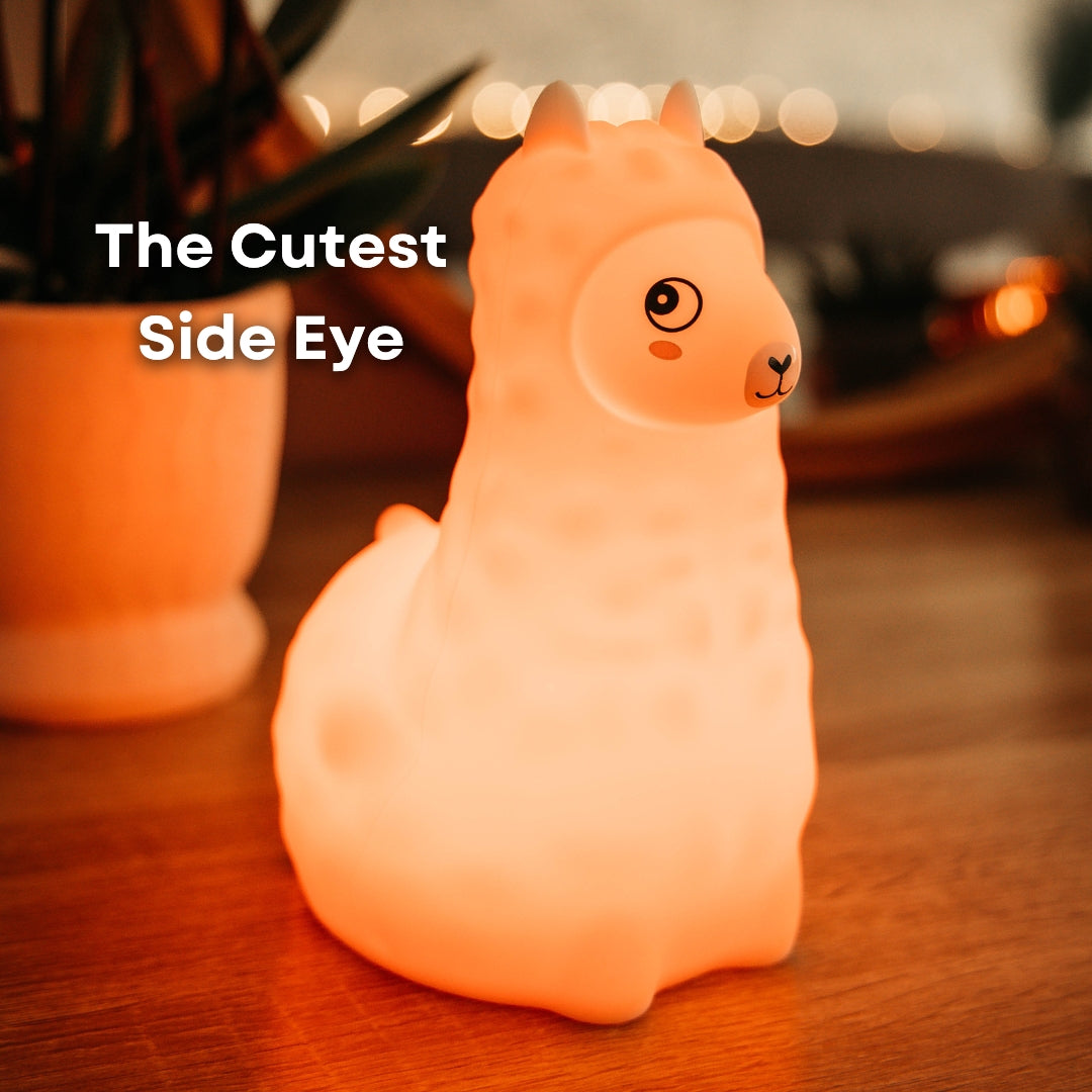 Lightie Llama silicone kids night light glowing warm white on a wooden surface with warm golden bokeh fairy lights in the background