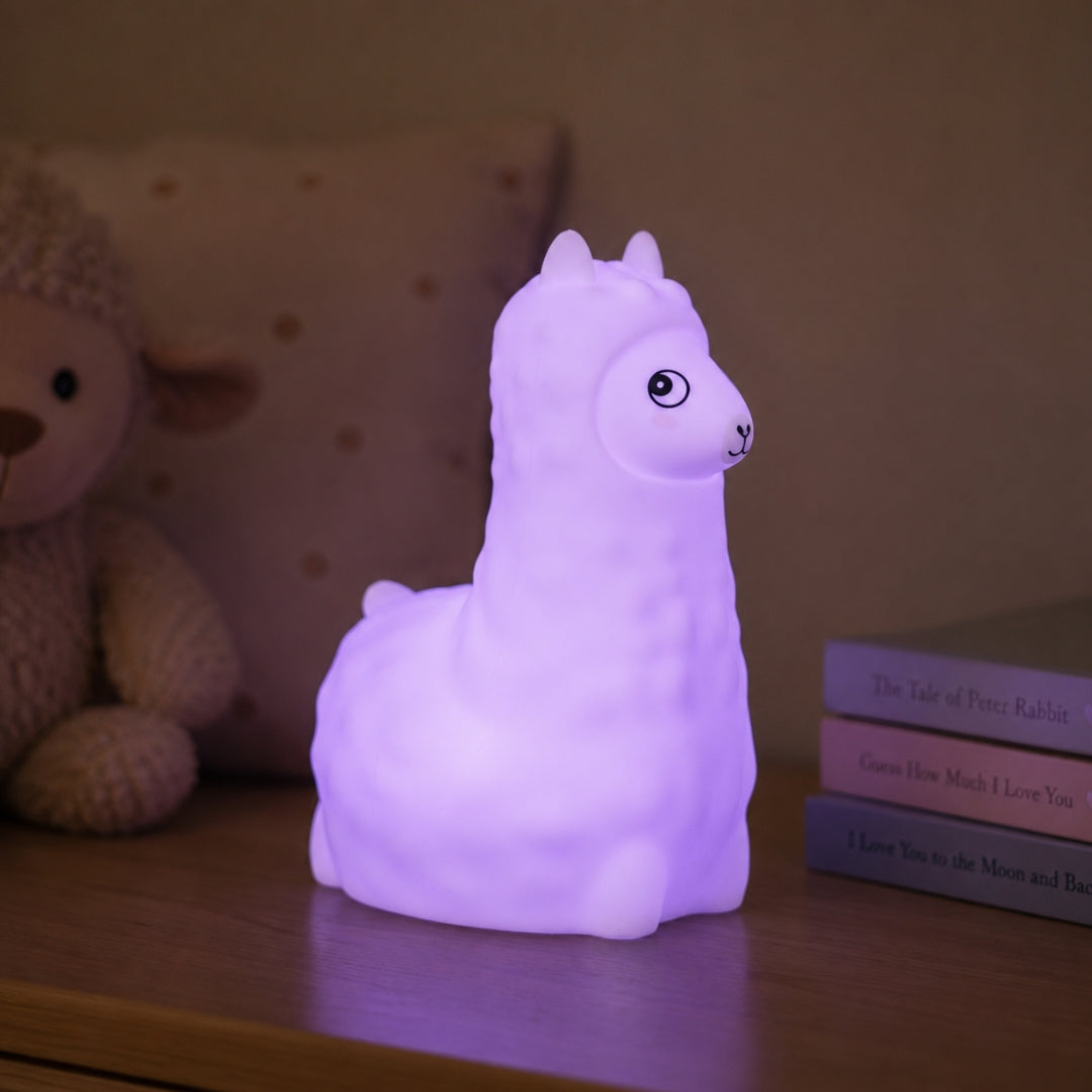 Lightie Llama silicone kids night light glowing warm white on a wooden bedside surface with cushions in the background