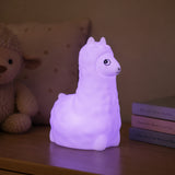 Lightie Llama silicone kids night light glowing warm white on a wooden bedside surface with cushions in the background