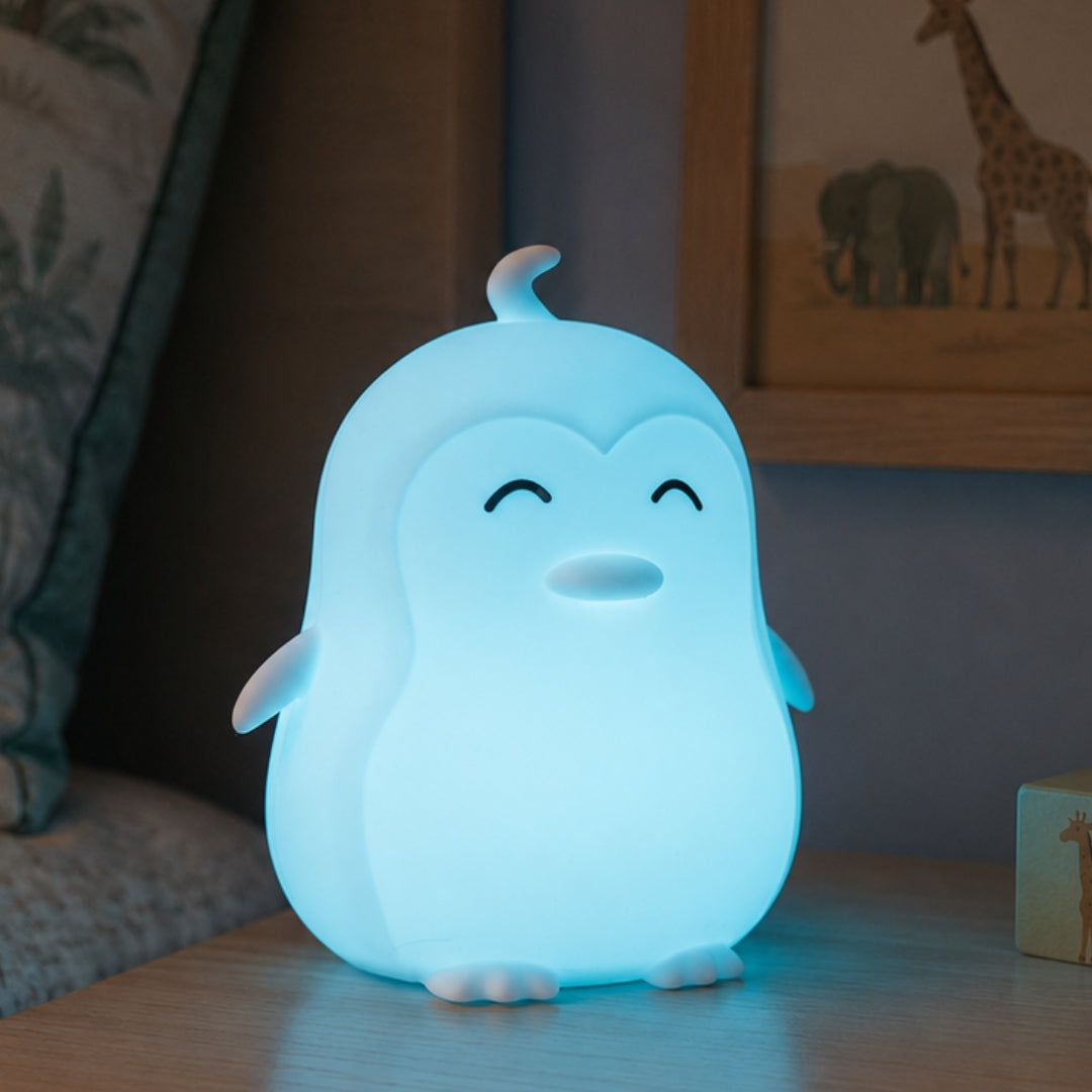 Lightie Penguin silicone kids night light glowing teal on a bedside table in a softly lit children's room with fairy lights in the background