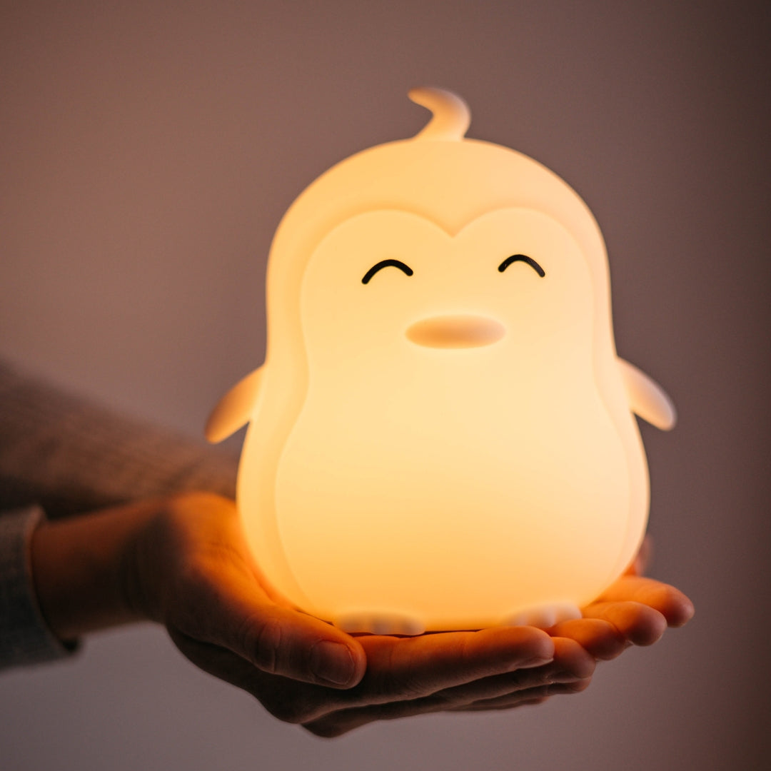 Lightie Penguin silicone kids night light glowing warm white on a wooden surface with warm golden bokeh fairy lights in the background
