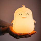 Lightie Penguin silicone kids night light glowing warm white on a wooden surface with warm golden bokeh fairy lights in the background
