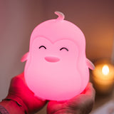 Lightie Penguin silicone kids night light glowing warm white on a wooden bedside surface with cushions in the background
