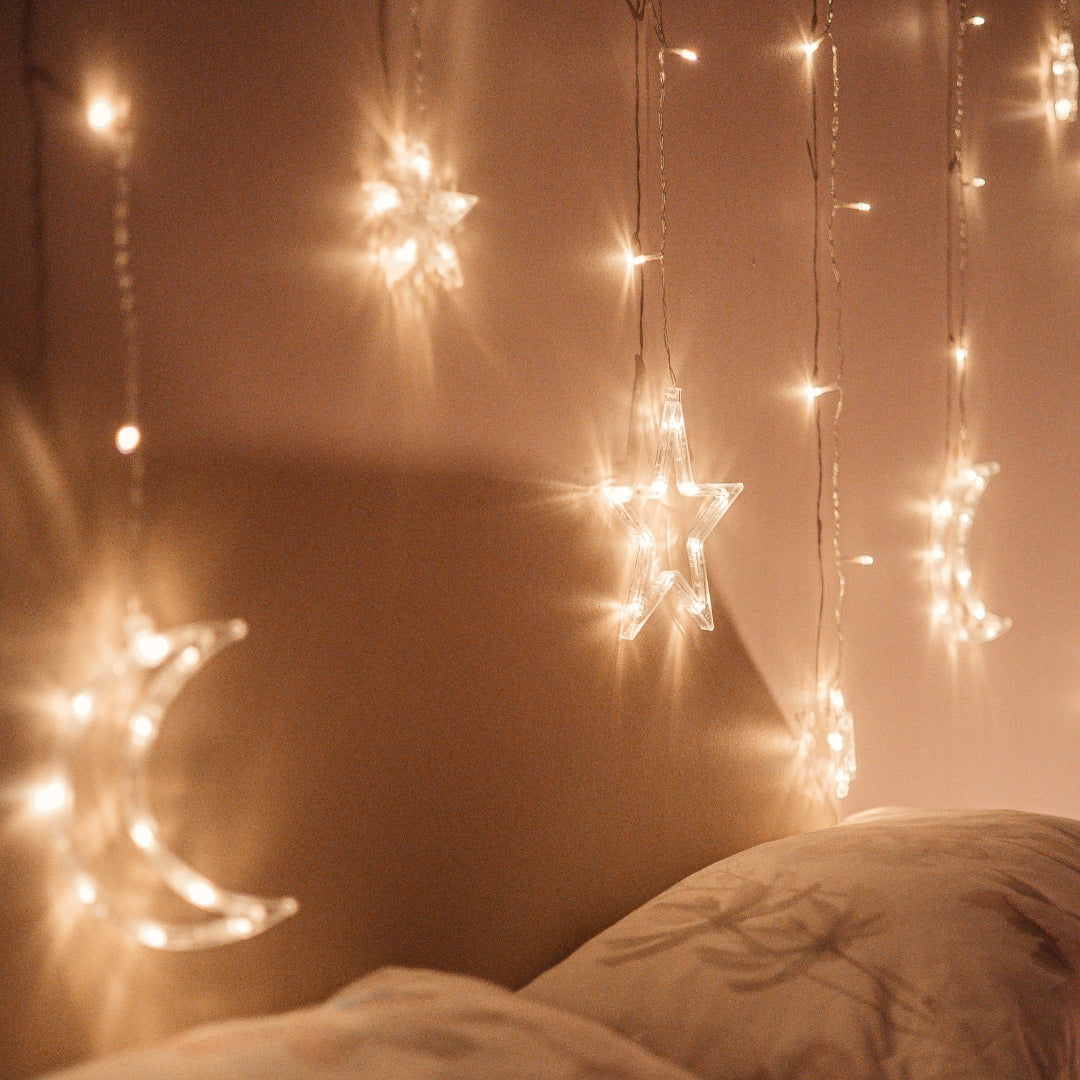 Moon and star curtain fairy lights draped above a bed against a bedroom wall