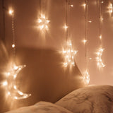 Moon and star curtain fairy lights draped above a bed against a bedroom wall