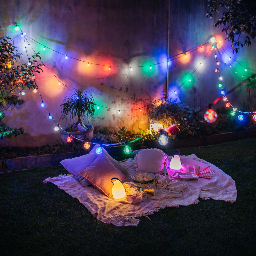 Outdoor backyard picnic setup at night with multicolour string lights draped over a blanket, pillows, lanterns, and food on grass.