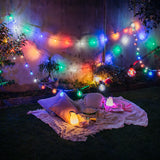 Outdoor backyard picnic setup at night with multicolour string lights draped over a blanket, pillows, lanterns, and food on grass.
