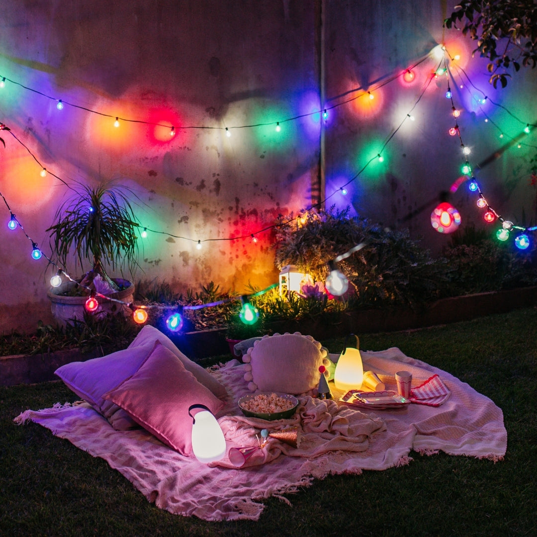 Multicolour outdoor string lights draped over a cozy picnic blanket on grass at night, with pillows, a glowing lamp, and a small lantern in a backyard setting.