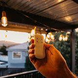 Hand holding the Festoon Remote On/Off Switch and Timer with Festoon string lights glowing overhead at dusk