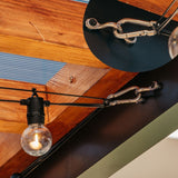 String Lighting suspended by black cords from silver metal hardware mounted on a dark beam, shown in a real interior setting with exposed wood ceiling panels.