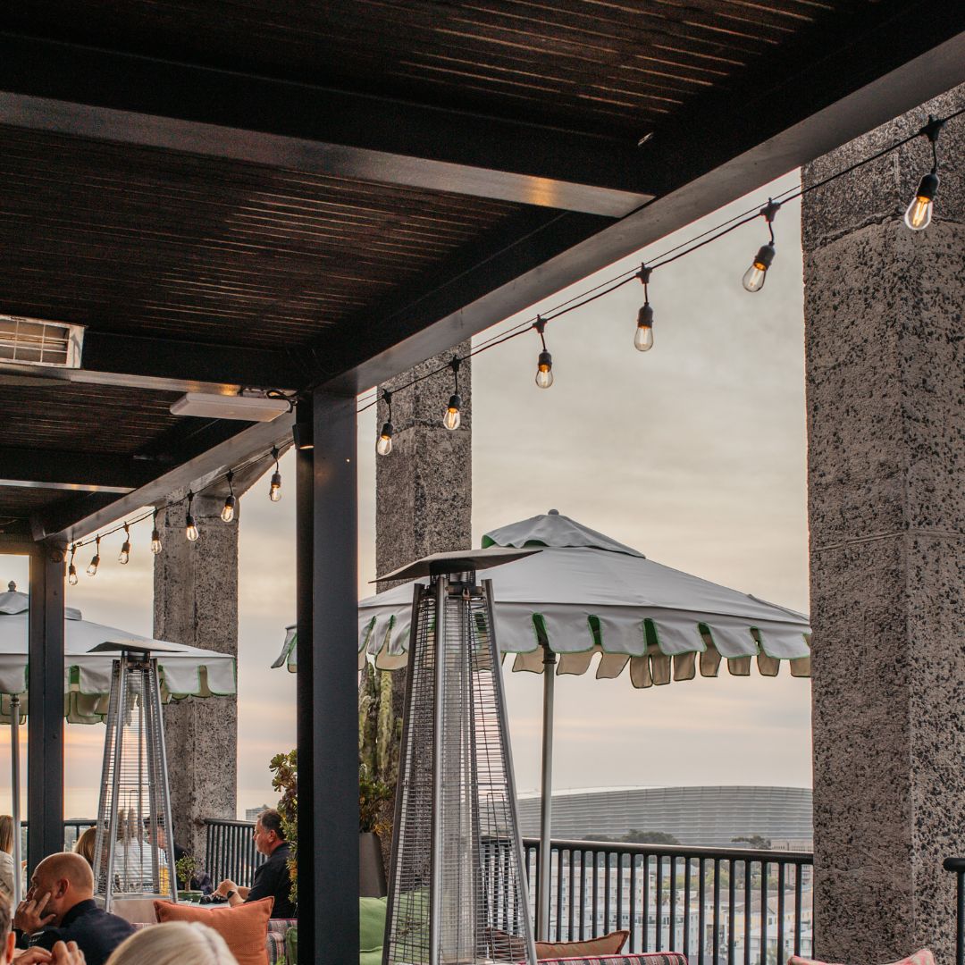 Outdoor patio seating area under a wooden pergola with hanging string lights, stone pillars, tall patio heaters, and large white umbrellas, with people seated in the foreground.