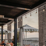 Outdoor patio seating area under a wooden pergola with hanging string lights, stone pillars, tall patio heaters, and large white umbrellas, with people seated in the foreground.