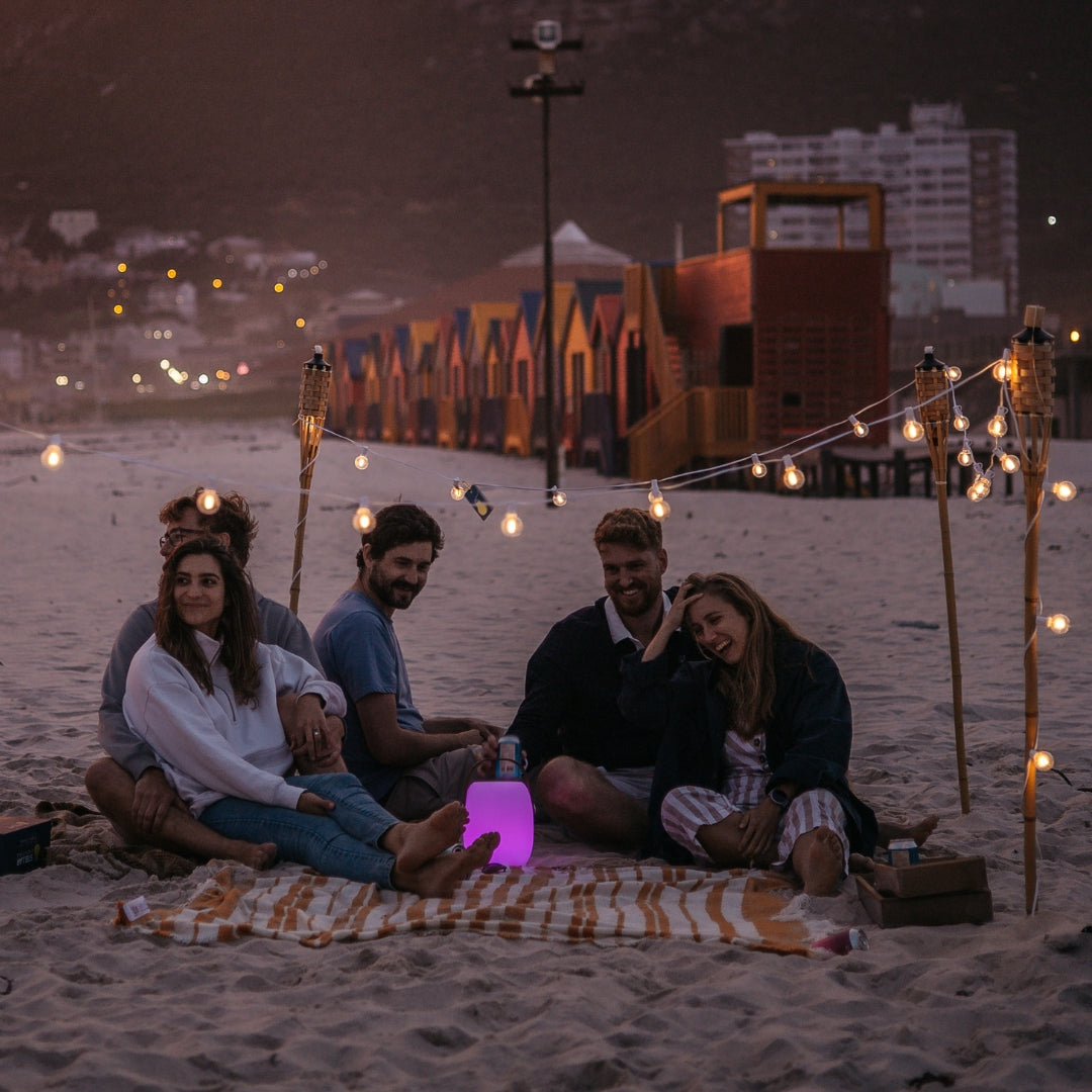 Group of friends sitting on a beach under solar string lights with G40 globe bulbs strung between bamboo poles at night with Muizenberg beach huts in the background