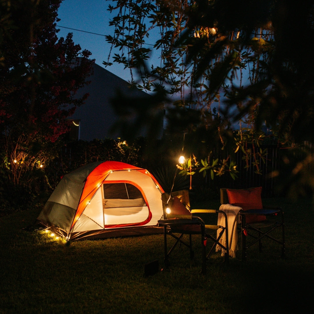 Solar string lights glowing around a camping tent with camp chairs in a dark garden at night