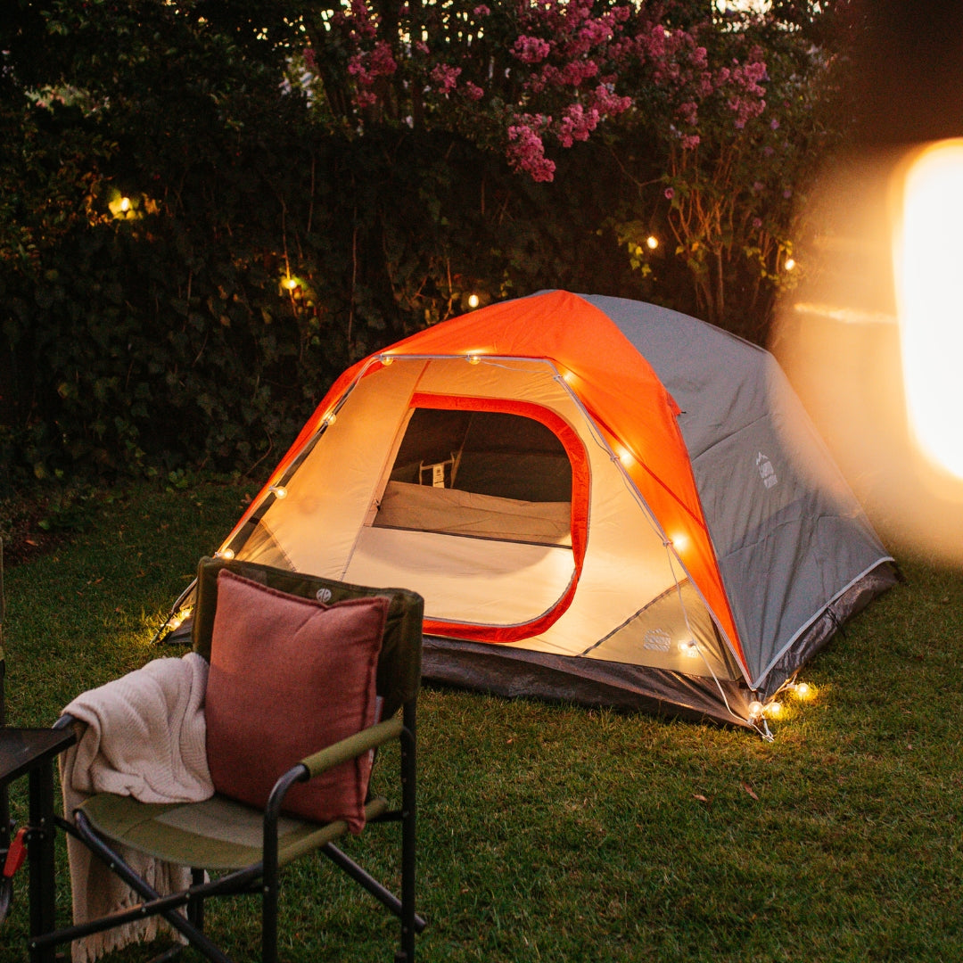 Solar string lights strung around a camping tent with camp chairs and blanket at dusk in a garden setting