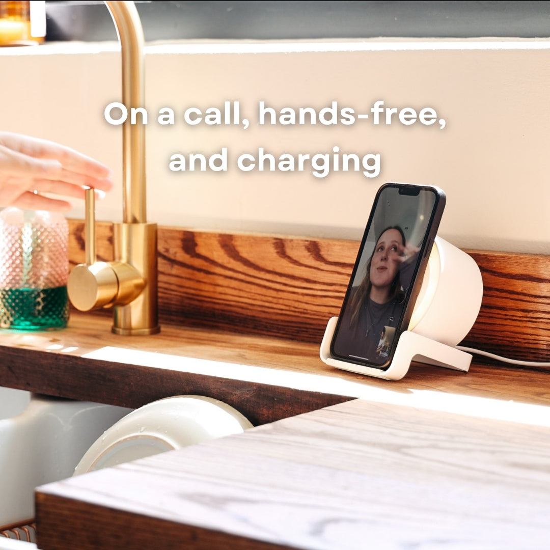 Smartphone propped on white speaker wireless charger during a video call on a kitchen counter beside a gold tap with hands-free and charging callout