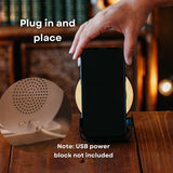 Hand placing smartphone onto white speaker wireless charger with inset close-up of rear speaker and on/off switch, note that USB power block is not included