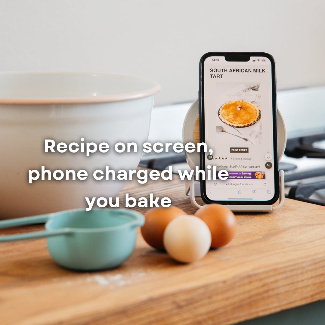 Smartphone displaying a South African milk tart recipe propped on bamboo speaker wireless charger on a kitchen counter beside eggs and mixing bowls