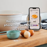 Smartphone displaying a South African milk tart recipe propped on bamboo speaker wireless charger on a kitchen counter beside eggs and mixing bowls
