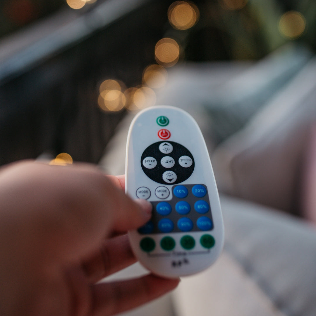 A hand holding a white multi-button remote control, photographed against a blurred outdoor background with warm bokeh lights.
