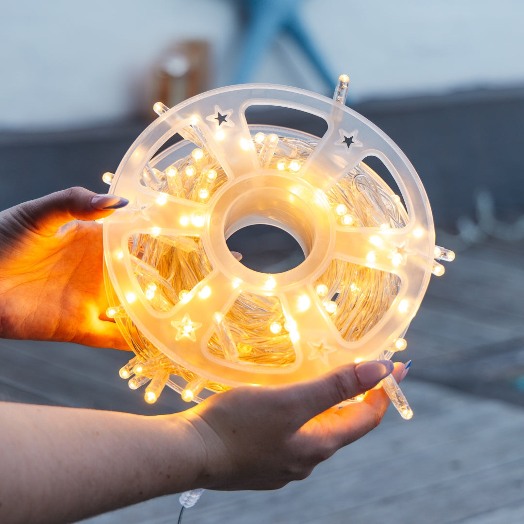 A person holds a large clear plastic spool of warm white fairy lights with LED bulbs, glowing brightly against an outdoor blurred background.