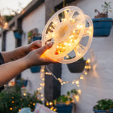 A person holds a translucent plastic spool of warm white LED fairy lights in their hands, with the lights glowing brightly. The background is a softly blurred outdoor patio setting with potted plants and more string lights hanging on a white wall.