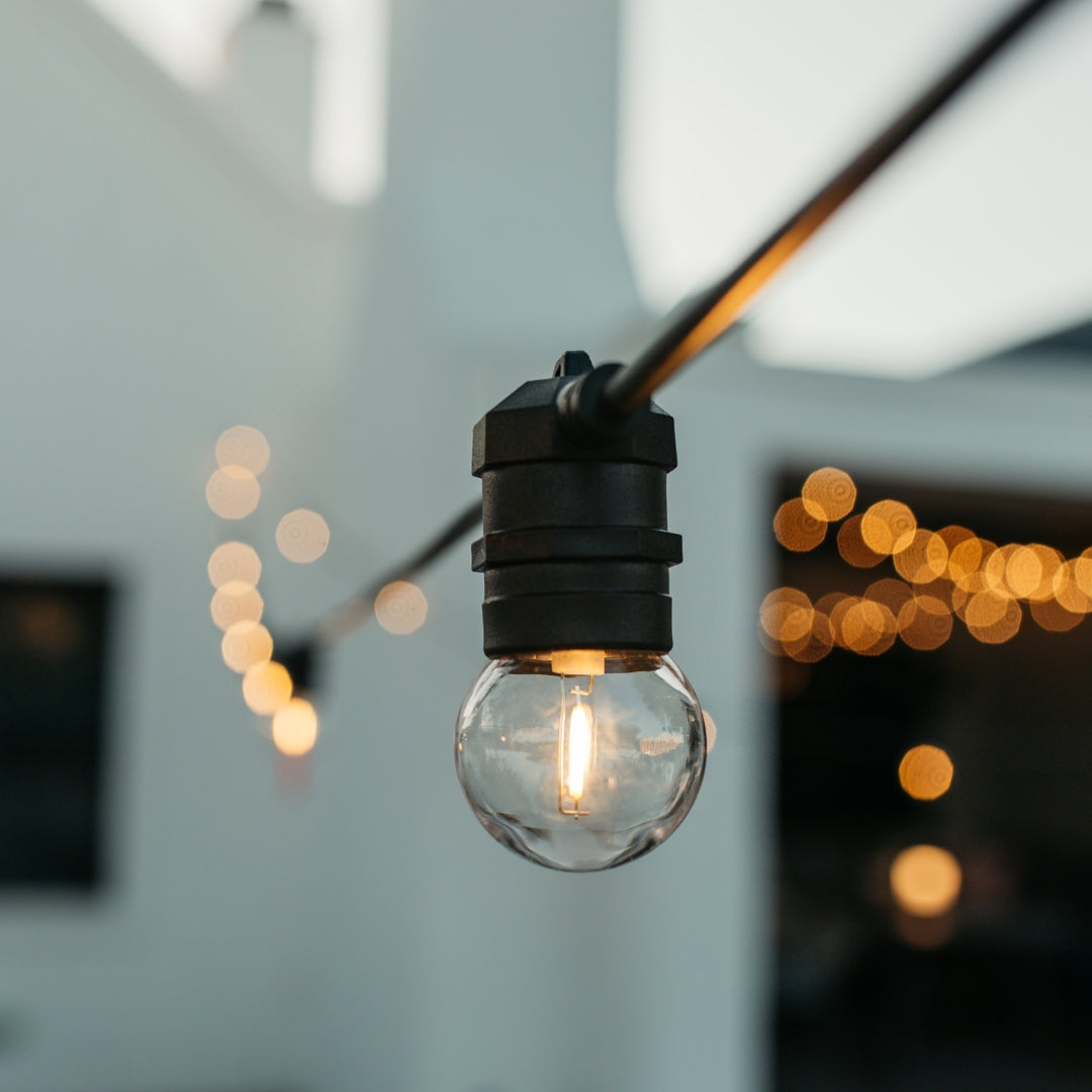 Close-up of warm white G45 retro globe bulb on black IP66 weatherproof festoon string lights against a white outdoor wall with warm bokeh background
