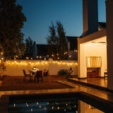 Ultimate Festoon String Lights strung across a South African garden at night with outdoor dining table set for dinner, braai area, and pool reflecting warm light in the foreground