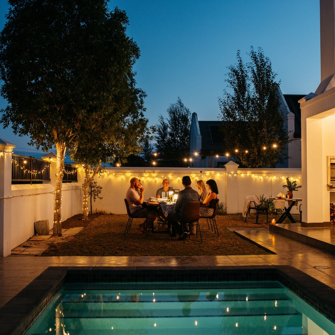 Group of friends dining outdoors at dusk under warm white Ultimate Festoon String Lights strung across a South African garden with a pool in the foreground