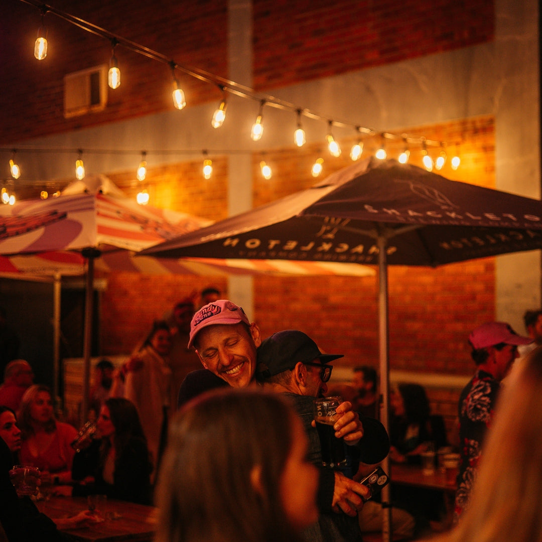 Two people hugging warmly at an outdoor evening event under Ultimate Gold Drop String Lights with warm golden T45 tubular bulbs suspended on individual drop wires creating a rich amber glow above