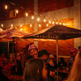 Two people hugging warmly at an outdoor evening event under Ultimate Gold Drop String Lights with warm golden T45 tubular bulbs suspended on individual drop wires creating a rich amber glow above