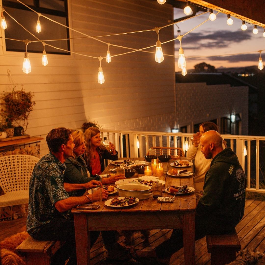 Group of friends dining outdoors at dusk under warm white Ultimate Drop String Lights suspended on individual drop wires in a South African garden