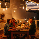 Group of friends dining outdoors at dusk under warm white Ultimate Drop String Lights suspended on individual drop wires in a South African garden
