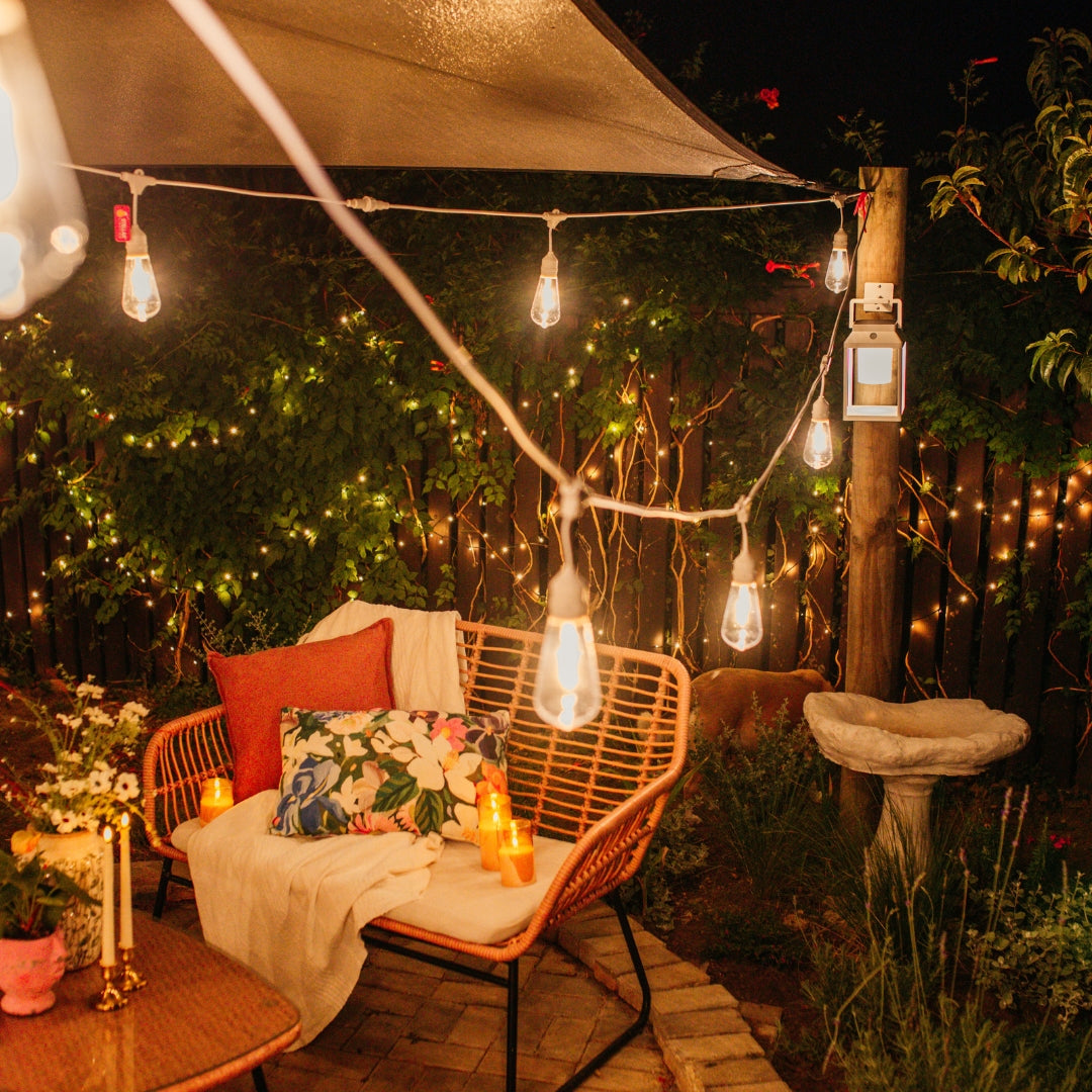 Styled South African outdoor patio at dusk showing Ultimate White Drop String Lights suspended on individual drop wires beneath a shade sail alongside fairy lights woven through hedges and pillar candles at ground level