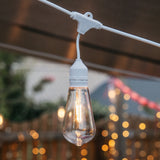 Close-up of warm white ST64 LED bulb suspended on individual drop wire on white IP66 weatherproof drop string lights against an outdoor background