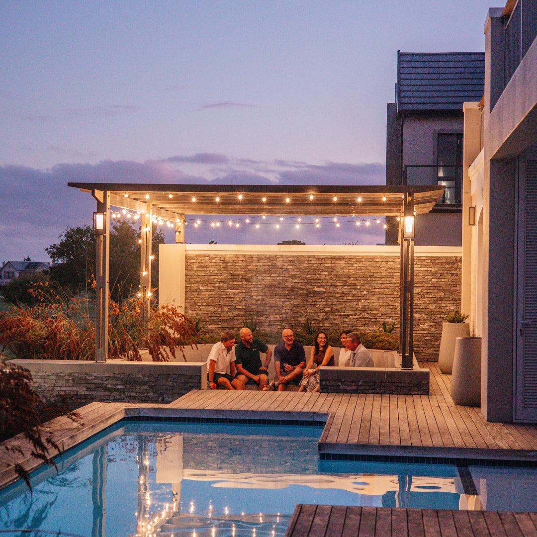 Group of friends gathered under vintage string lights on a pergola beside a pool at dusk