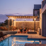 Group of friends gathered under vintage string lights on a pergola beside a pool at dusk