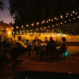 Group of friends dining outdoors under warm amber vintage string lights strung across a garden patio at night