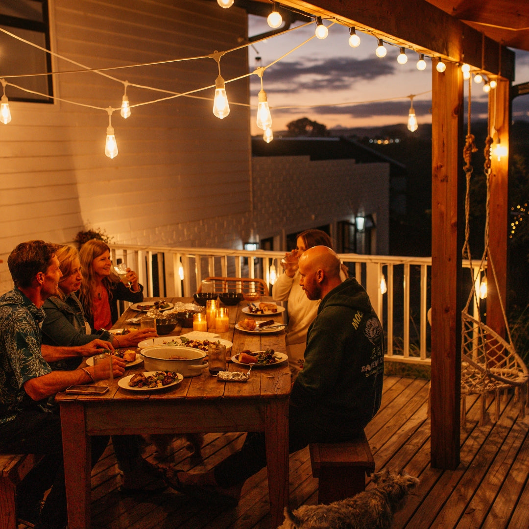 Group of people dining on a wooden deck with string lights and a sunset view.