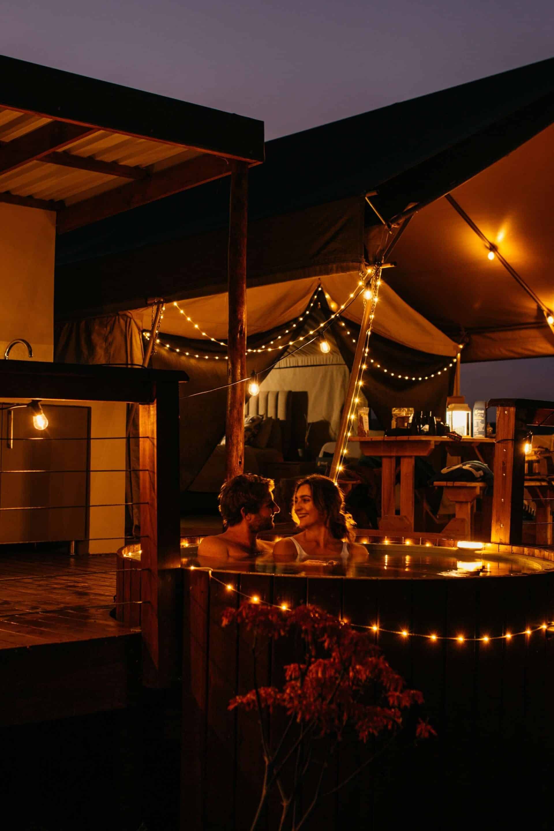 Couple relaxing in a wood-fired hot tub beside a glamping tent at Longhill Reserve, lit with warm solar string lights