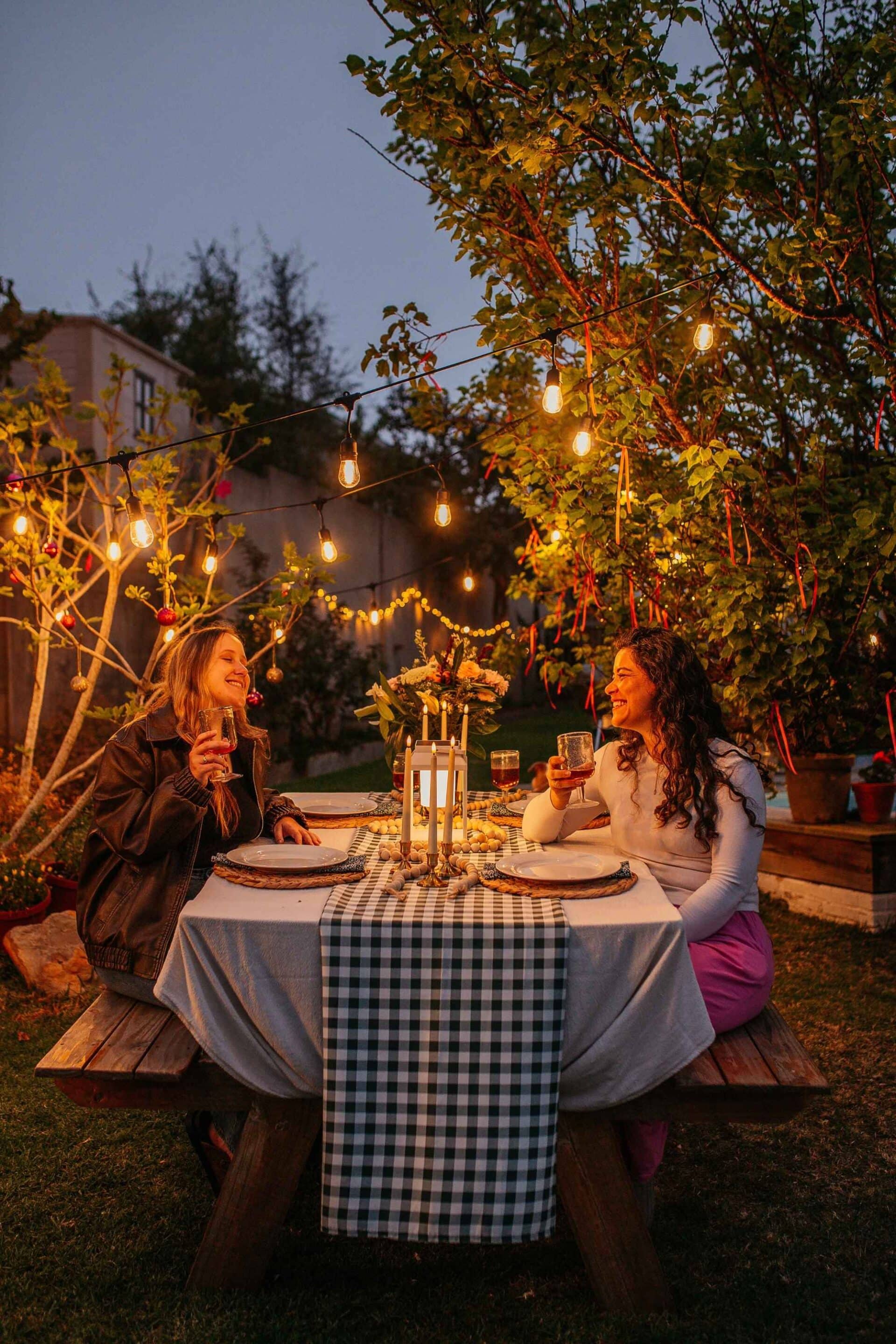 Two women enjoying a candlelit garden dinner under festoon drop string lights